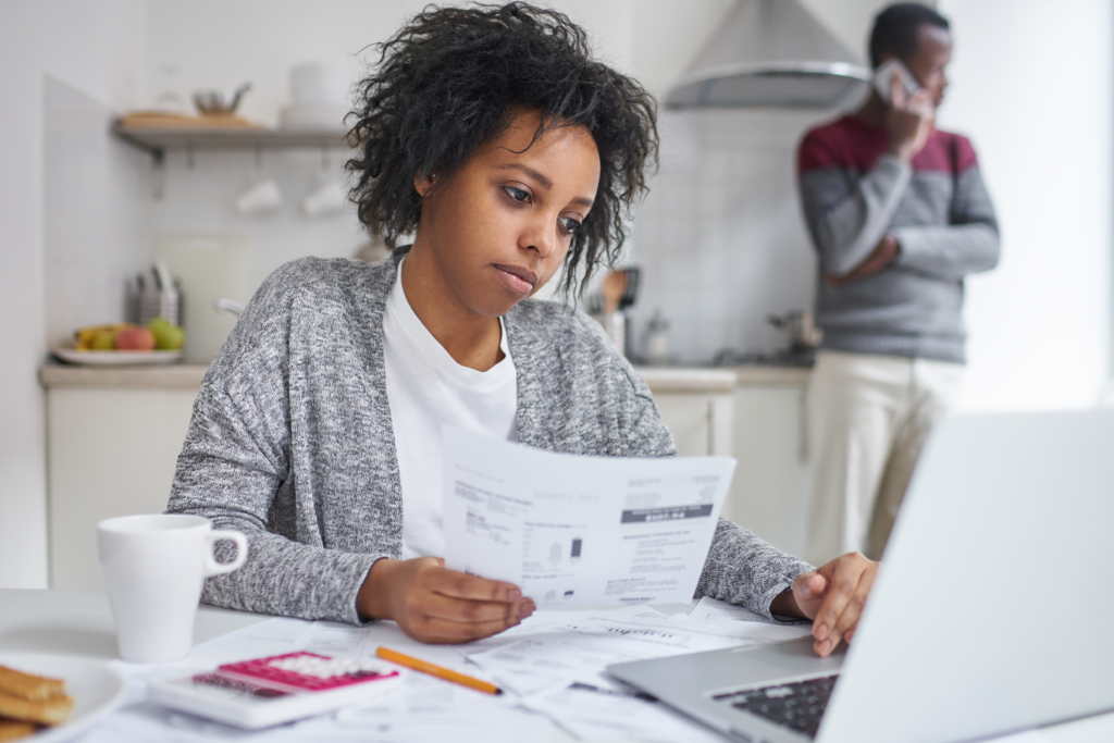 Woman reviewing bills at a table with a laptop while another person stands in the background on the phone.