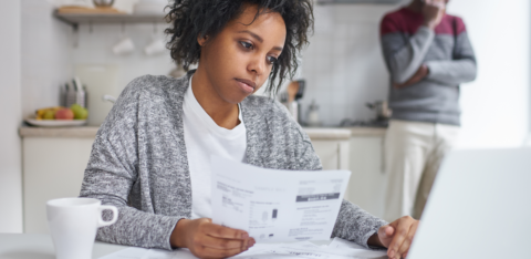 Woman reviewing bills at a table with a laptop while another person stands in the background on the phone.