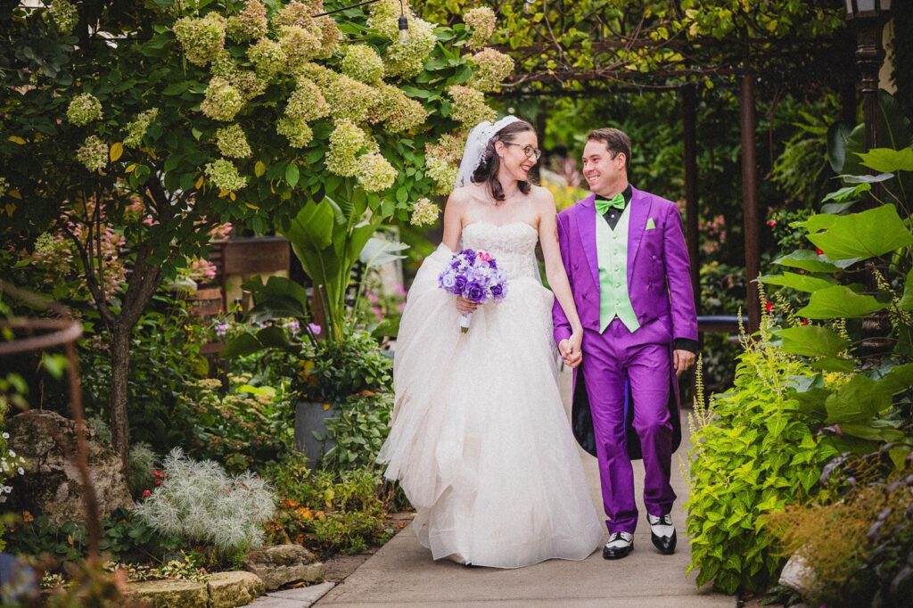 Bride and groom walking hand in hand through a garden, smiling at each other.