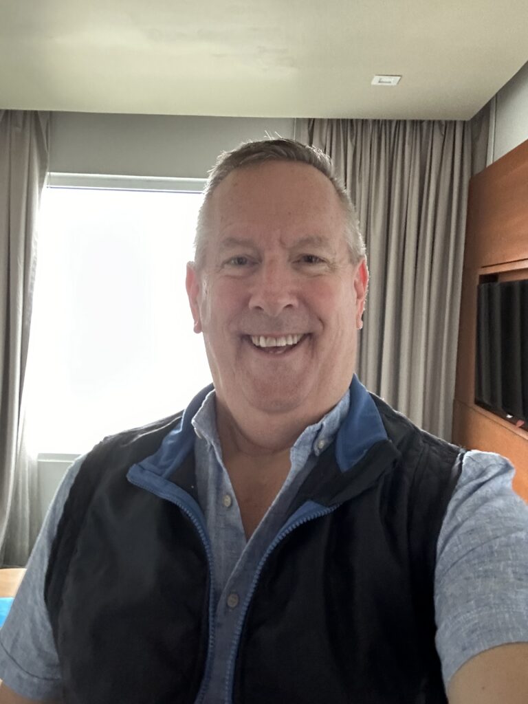 Smiling man taking a selfie indoors with a window and curtains in the background.