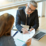 Man in a suit reviewing documents with a client at a desk, pointing to a chart on the paper.