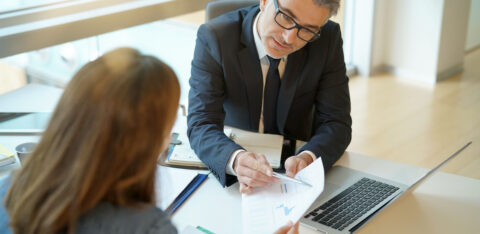 Man in a suit reviewing documents with a client at a desk, pointing to a chart on the paper.