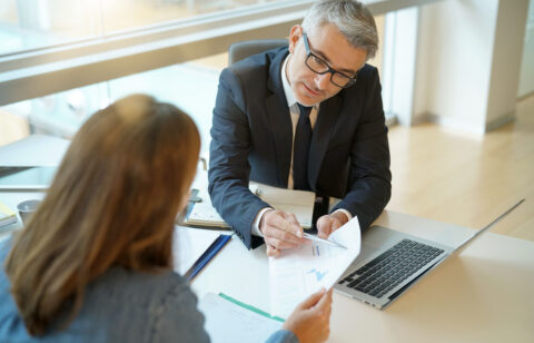 Man in a suit reviewing documents with a client at a desk, pointing to a chart on the paper.