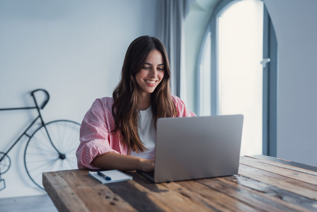 Person smiling while using a laptop at a wooden table, with a notebook nearby