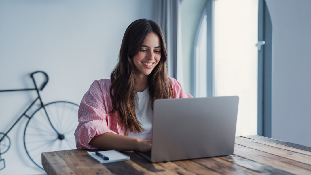 Person smiling while using a laptop at a wooden table, with a notebook nearby