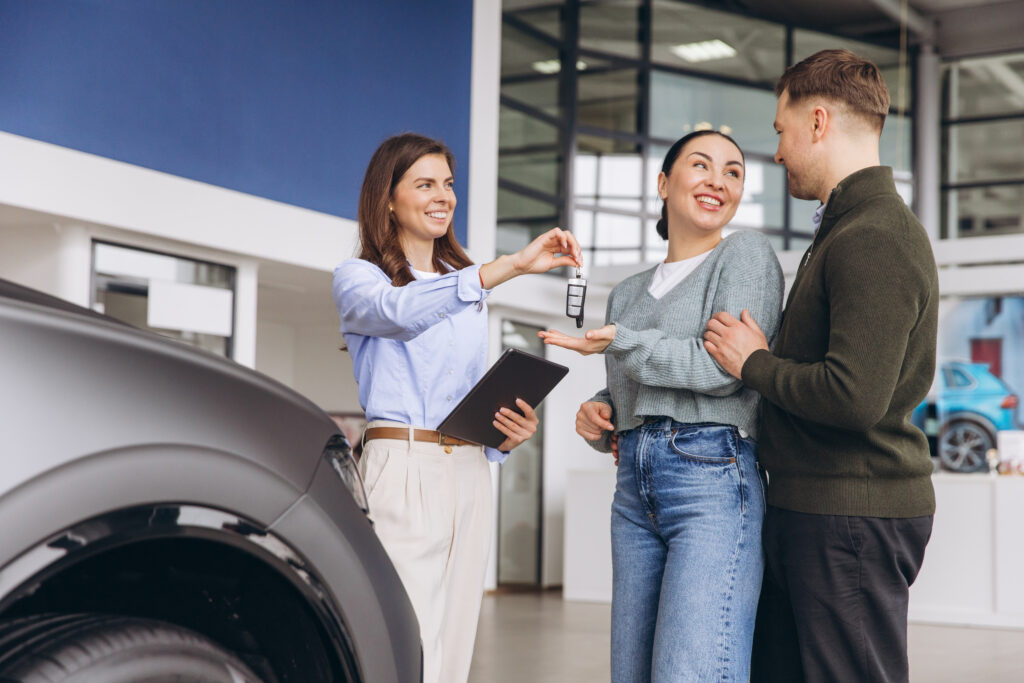 Saleswoman handing Salesperson handing car keys to a smiling couple at a dealership, with a vehicle nearby.