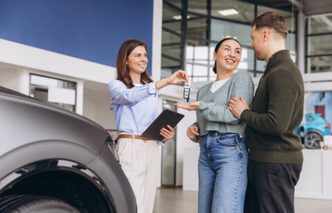 Saleswoman handing Salesperson handing car keys to a smiling couple at a dealership, with a vehicle nearby.