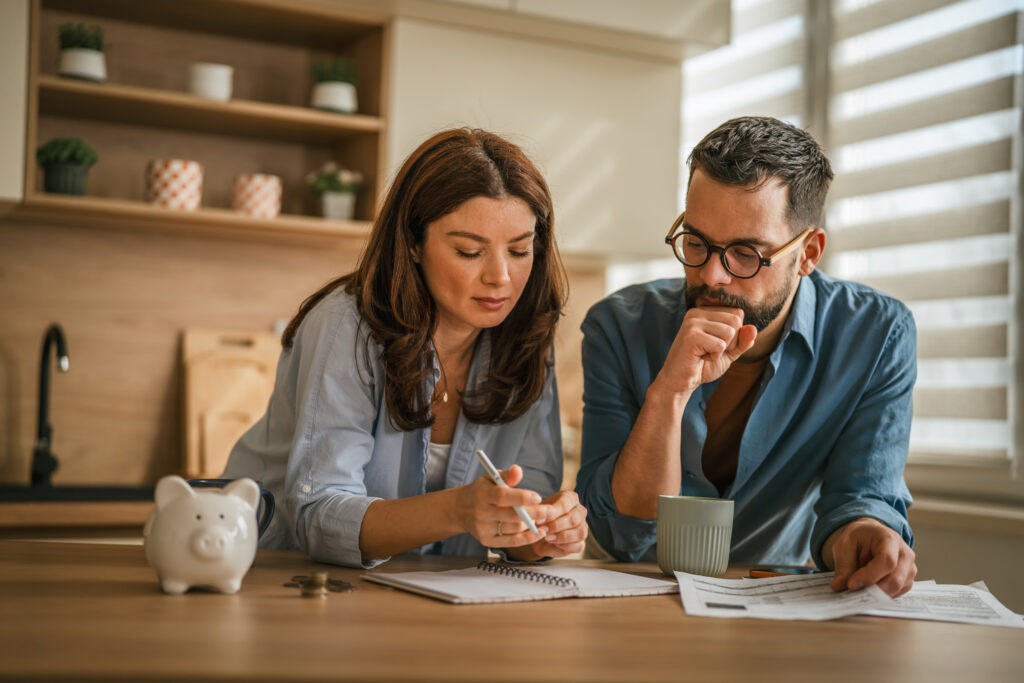 Two adults looking over financial documents and taking notes at home, with coins and a piggy bank on the table.