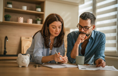 Two adults looking over financial documents and taking notes at home, with coins and a piggy bank on the table.