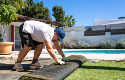 Middle aged man removing artificial grass in a sunny backyard with a swimming pool, showing residential landscaping work, lawn replacement and outdoor home improvement project