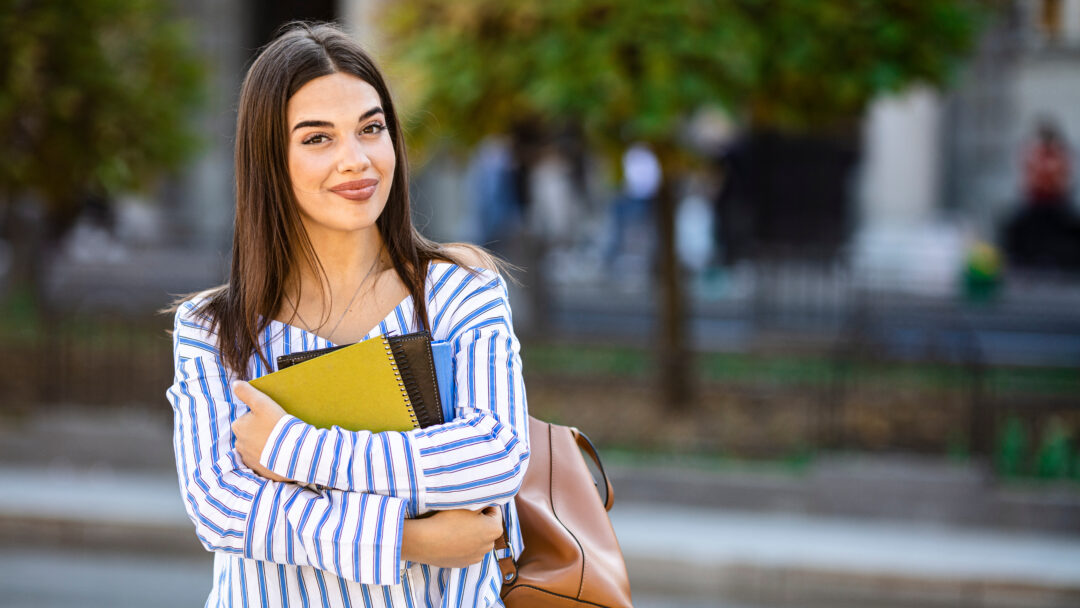 Student carrying books and a shoulder bag, standing on a city street with trees in the background.