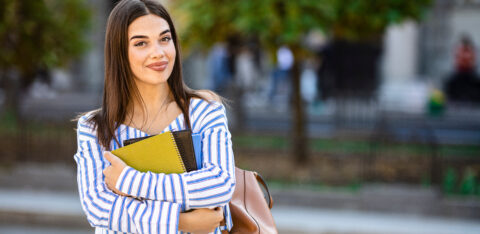 Student carrying books and a shoulder bag, standing on a city street with trees in the background.