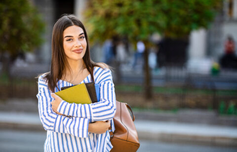 Student carrying books and a shoulder bag, standing on a city street with trees in the background.