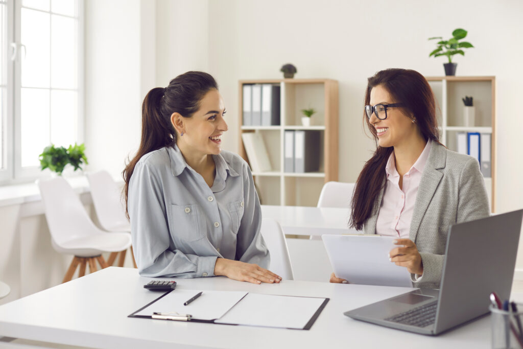 Two women sitting at a desk in an office, smiling and reviewing documents together.