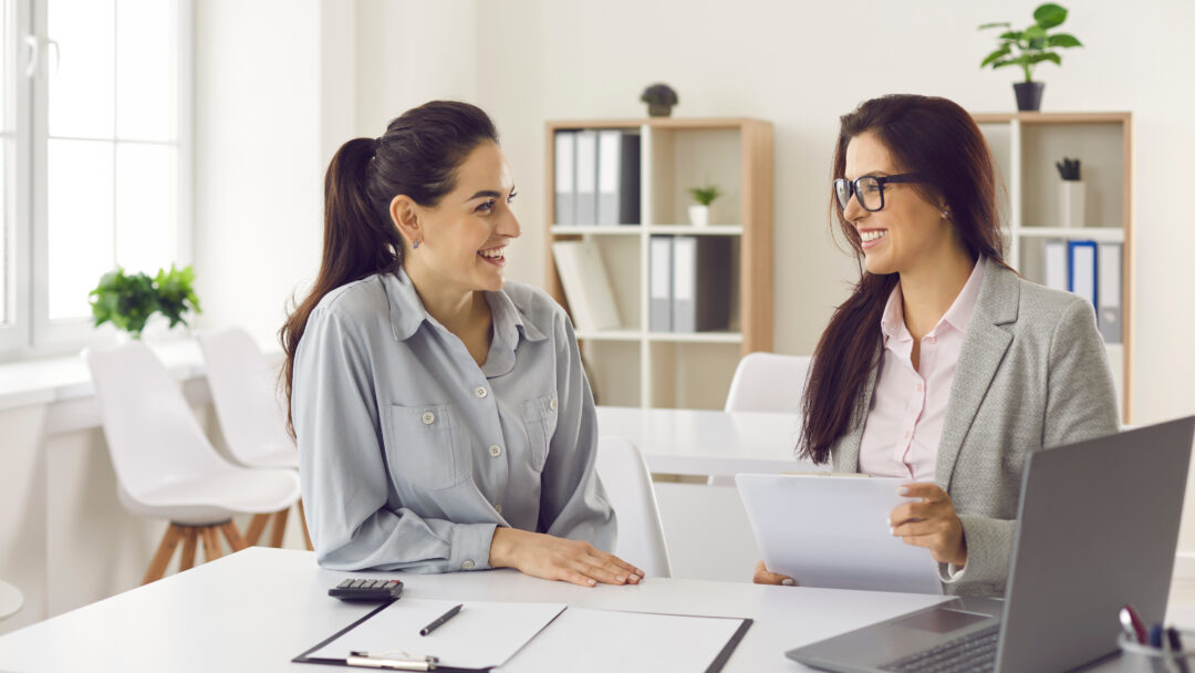 Two women sitting at a desk in an office, smiling and reviewing documents together.