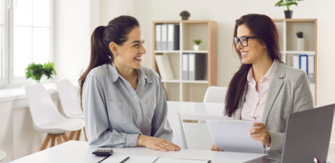 Two women sitting at a desk in an office, smiling and reviewing documents together.