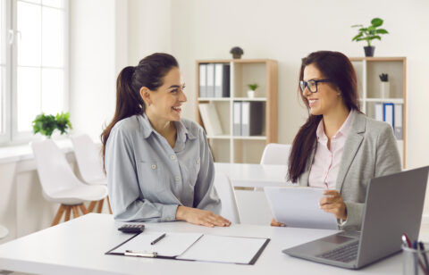 Two women sitting at a desk in an office, smiling and reviewing documents together.