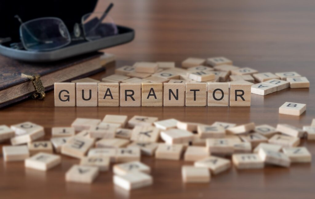 Wooden letter tiles spelling “guarantor” on a table, with scattered tiles and books in the background.