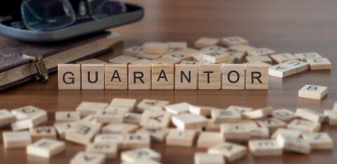 Wooden letter tiles spelling “guarantor” on a table, with scattered tiles and books in the background.