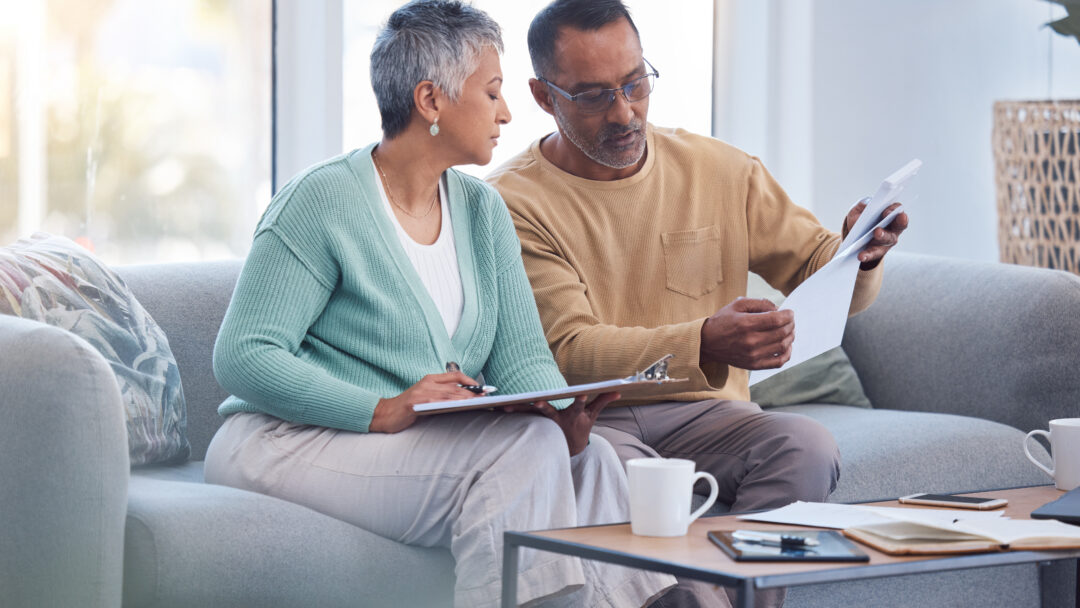 Older couple reviewing paperwork together on a couch, with one holding a document and the other taking notes