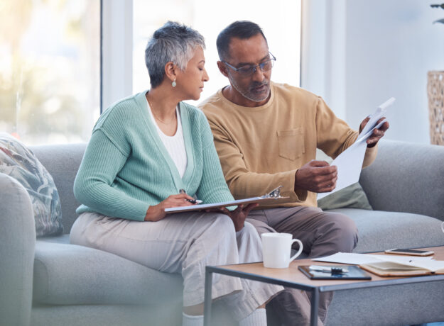 Older couple reviewing paperwork together on a couch, with one holding a document and the other taking notes