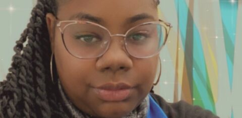 Woman with braided hair and glasses taking a selfie indoors, wearing a scarf and blue shirt