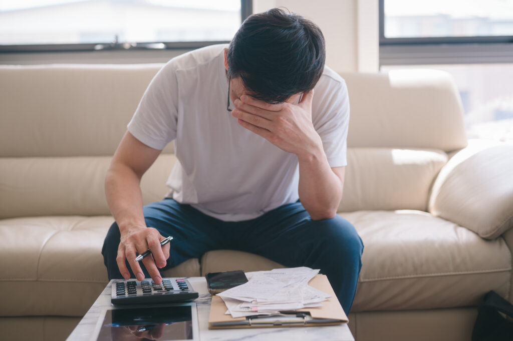 Person sitting on a couch reviewing bills, using a calculator while covering their face in stress