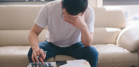Person sitting on a couch reviewing bills, using a calculator while covering their face in stress