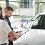 Customer signing paperwork on a clipboard while standing next to a car with a salesperson