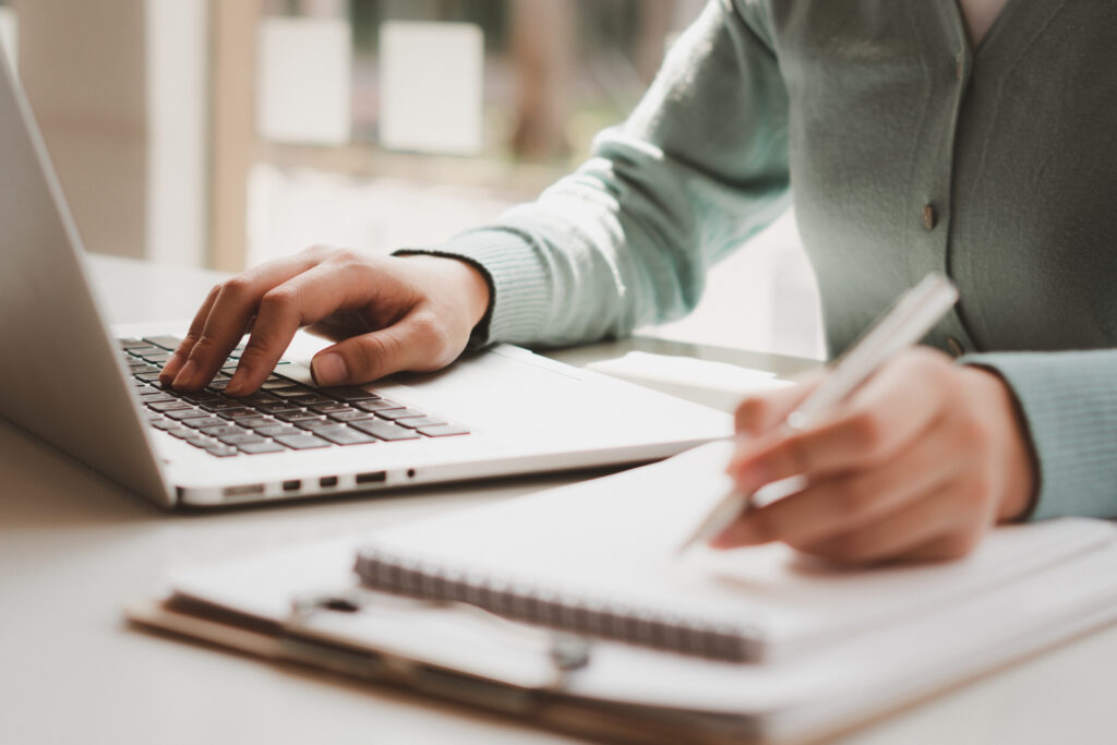 Close-up of hands typing on a laptop while writing notes in a notebook