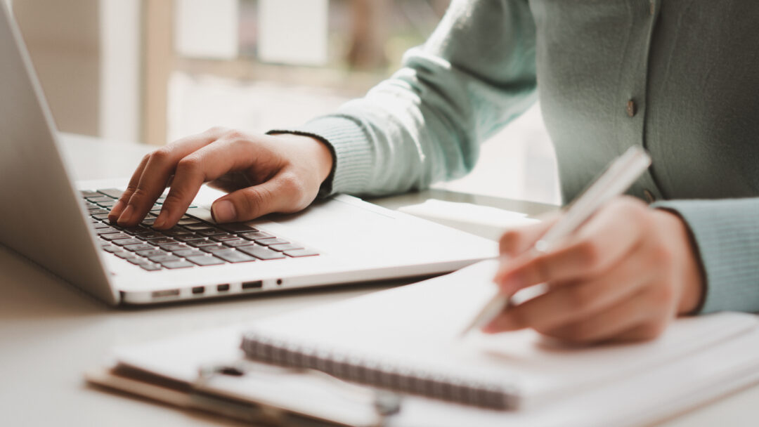 Close-up of hands typing on a laptop while writing notes in a notebook
