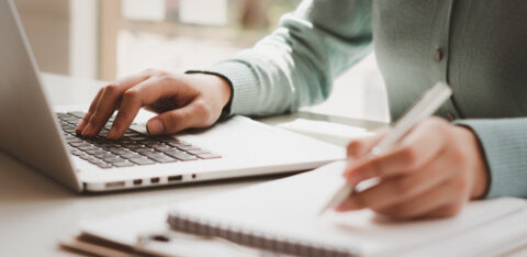 Close-up of hands typing on a laptop while writing notes in a notebook
