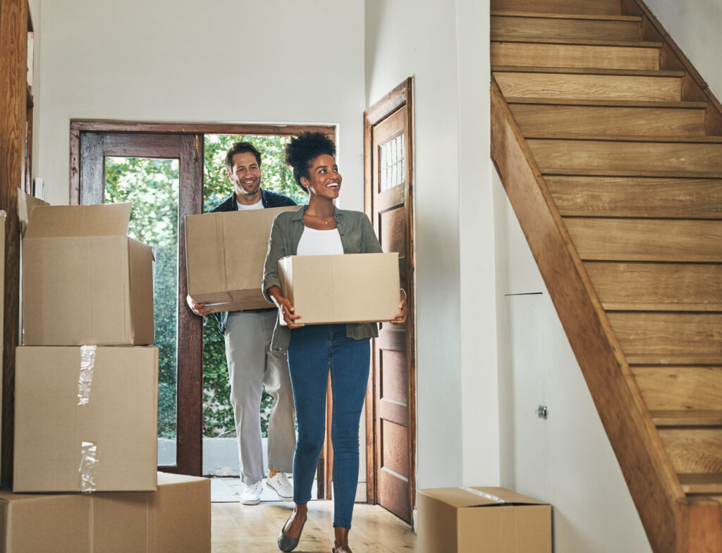 Couple carrying moving boxes into a home, stepping through the front door with more boxes nearby