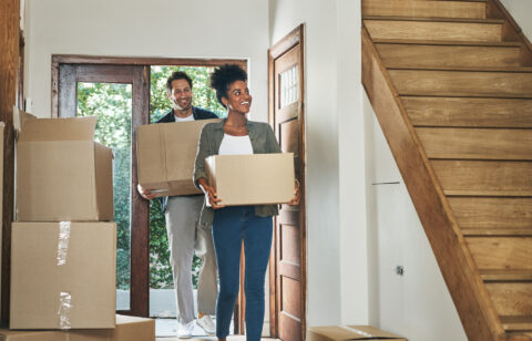 Couple carrying moving boxes into a home, stepping through the front door with more boxes nearby